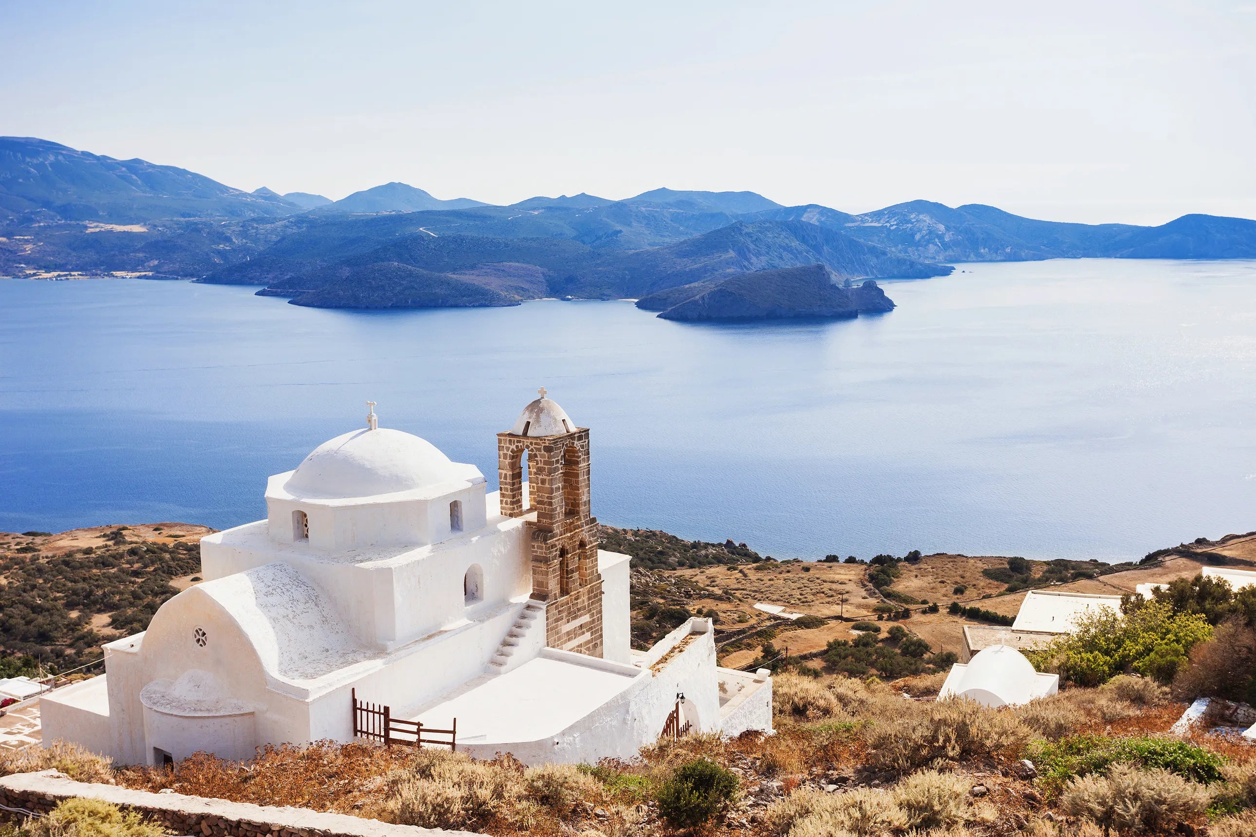 An iconic white building in a greek island with an astonishing water-side view with some mountains in the back